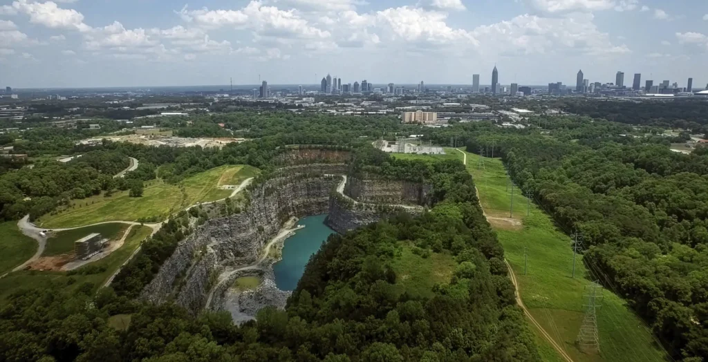 Aerial view of old quarry filled with water surrounded by grass and trees with a city in the background